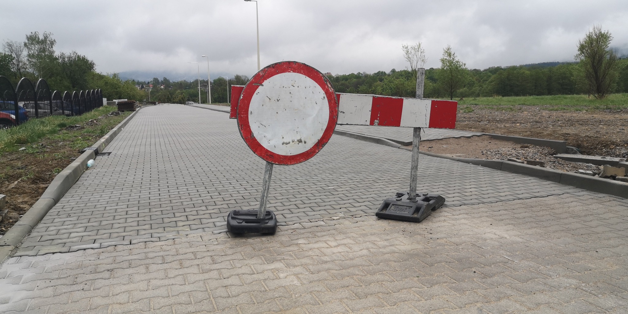Świeżo ułożona szara kostka brukowa z widocznym znakiem zakazu wjazdu i barierami drogowymi, w tle zieleń i góry w pochmurny dzień.