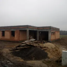 Surowy stan domu z czerwonej cegły, widoczne niezadaszone okna, betonowy wieniec, sterta desek i żwiru na błotnistym placu budowy w pochmurny dzień.