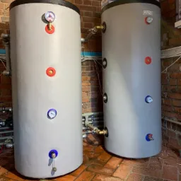 Dwa szare zbiorniki akumulacyjne ciepła z manometrami i zaworami, połączone rurami miedzianymi, ustawione w pomieszczeniu z ceglanymi ścianami i terakotową podłogą.