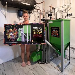 Słoneczny Doraca PL Sp. z o.o. - Kobieta prezentuje voucher na kocioł na pellet 18kW z instalacją, stojąc obok kotła. Widoczne logo firmy słonecznydoradca.pl i informacja o terminie ważności vouchera.