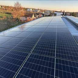 Słoneczny Doraca PL Sp. z o.o. - Instalacja paneli słonecznych na dachu budynku mieszkalnego, widok z góry, osoba na dachu, w tle zabudowa jednorodzinna i drzewa, słoneczny dzień.