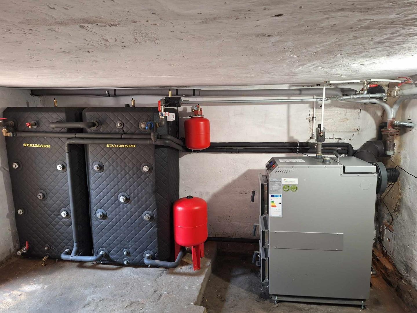 Szary piec na paliwo stałe Stalmark z dwoma czarnymi zbiornikami akumulacyjnymi w kotłowni. Widoczne rury i czerwone zbiorniki wyrównawcze.