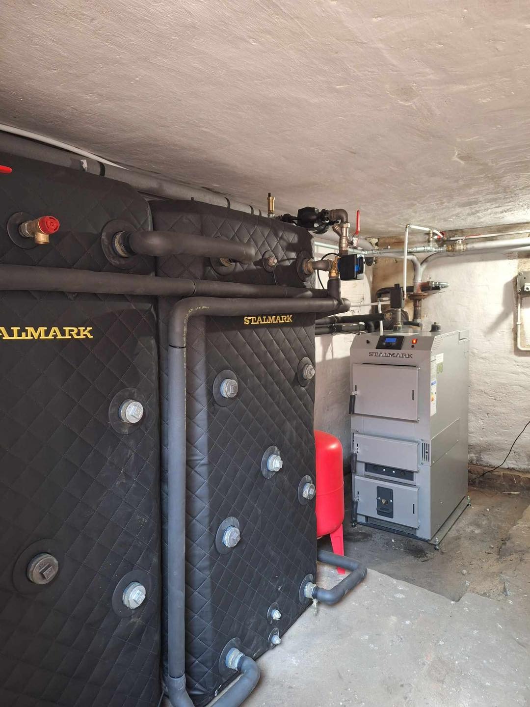 Kocioł na paliwo stałe marki Stalmark z czarną, pikowaną obudową i instalacją hydrauliczną, widoczny w kotłowni. Ujęcie z niskiego kąta.