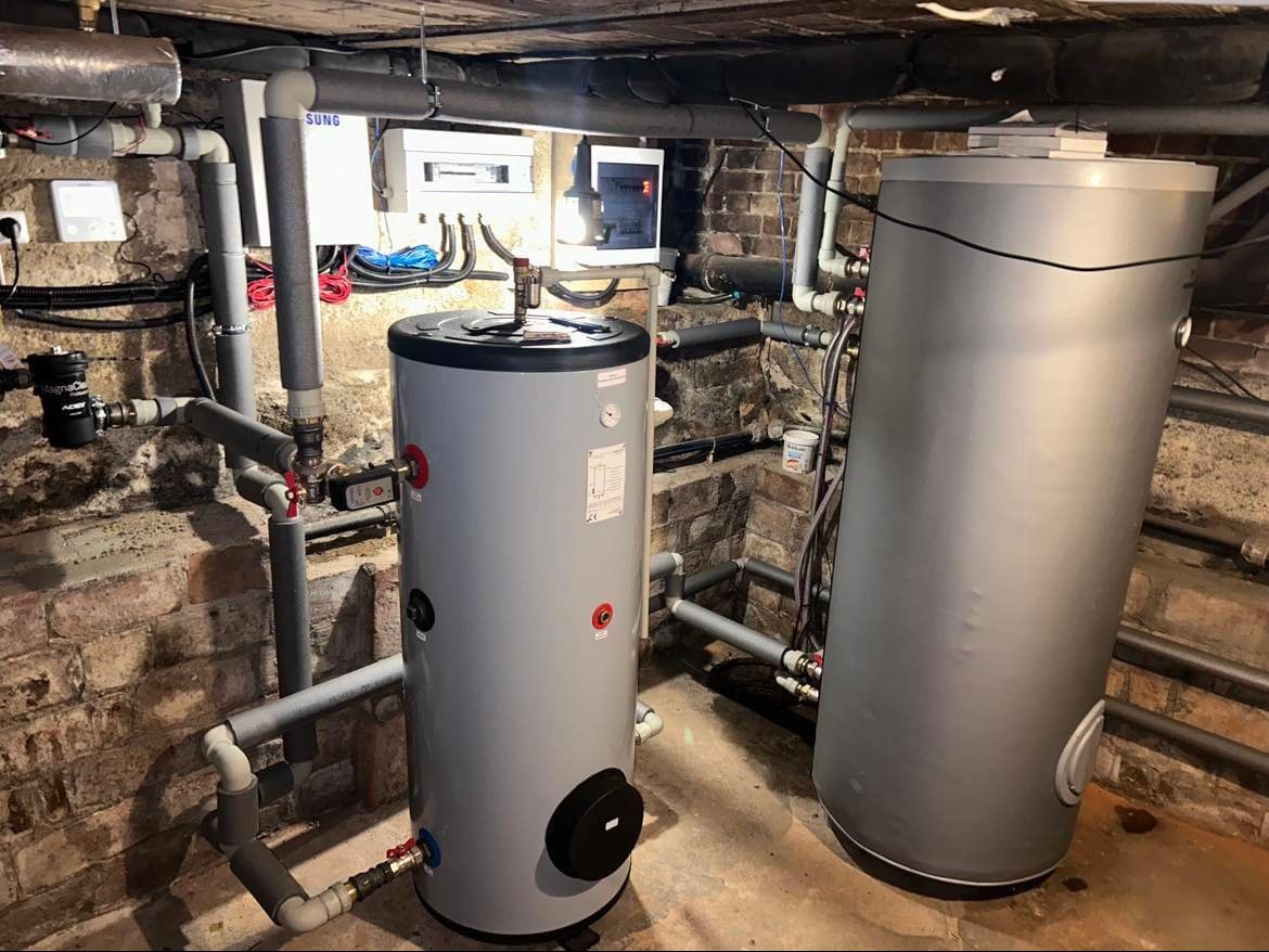 Dwa zbiorniki akumulacyjne w kotłowni z ceglanymi ścianami i szarymi rurami, instalacja w Ostrowie Wielkopolskim. Widoczne elementy sterowania i zabezpieczeń.