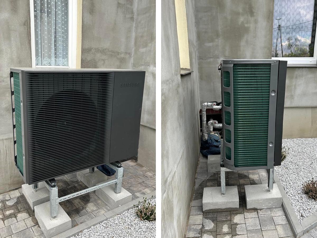 Czarna pompa ciepła Samsung na betonowych podstawach, zamontowana przy szarej ścianie budynku z oknem. Widoczne rury i kostka brukowa.