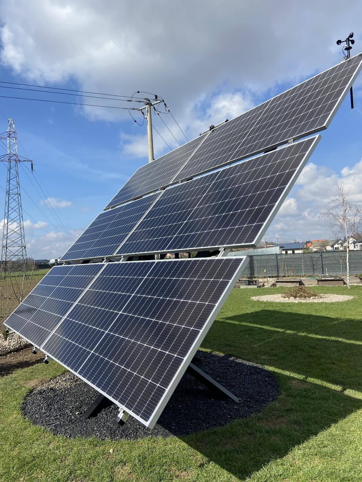 Instalacja paneli fotowoltaicznych na trawniku, z widoczną linią energetyczną i wiatromierzem. Panele ustawione pod kątem, otoczone czarnym żwirem.