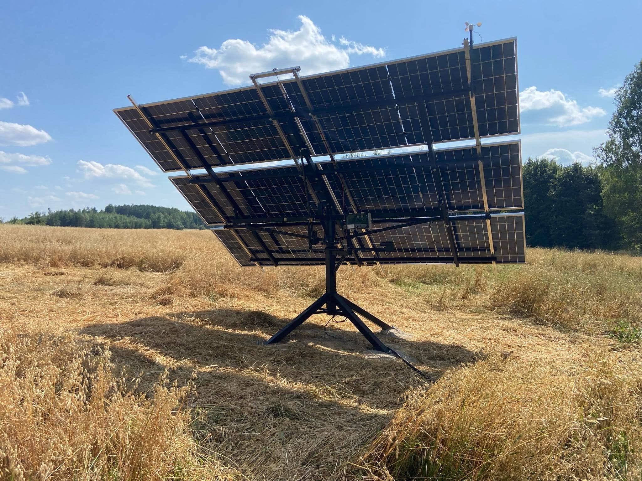 Dwu-panelowa instalacja solarna na trójnożnej podstawie, umieszczona na polu ze złotą trawą, pod błękitnym niebem z chmurami. Widok z tyłu paneli.