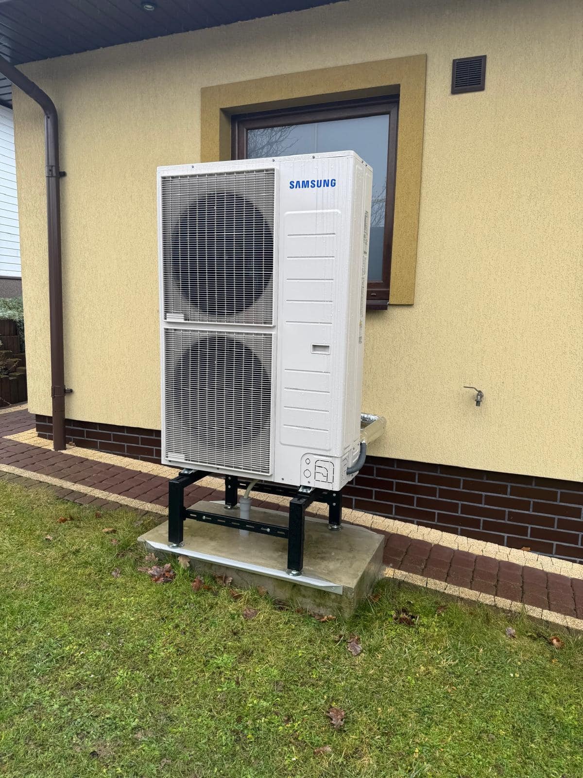 Biała pompa ciepła Samsung, dwusekcyjna, zamontowana na czarnej, metalowej konstrukcji na betonowej podstawie, przy żółtej ścianie budynku z brązowymi detalami.