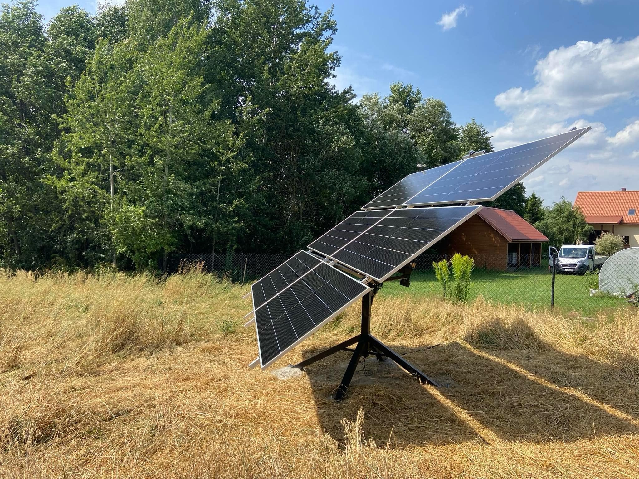 Mobilny zestaw paneli fotowoltaicznych na trójnogu, ustawiony na wyschniętej trawie, z domem i drzewami w tle, w słoneczny dzień.