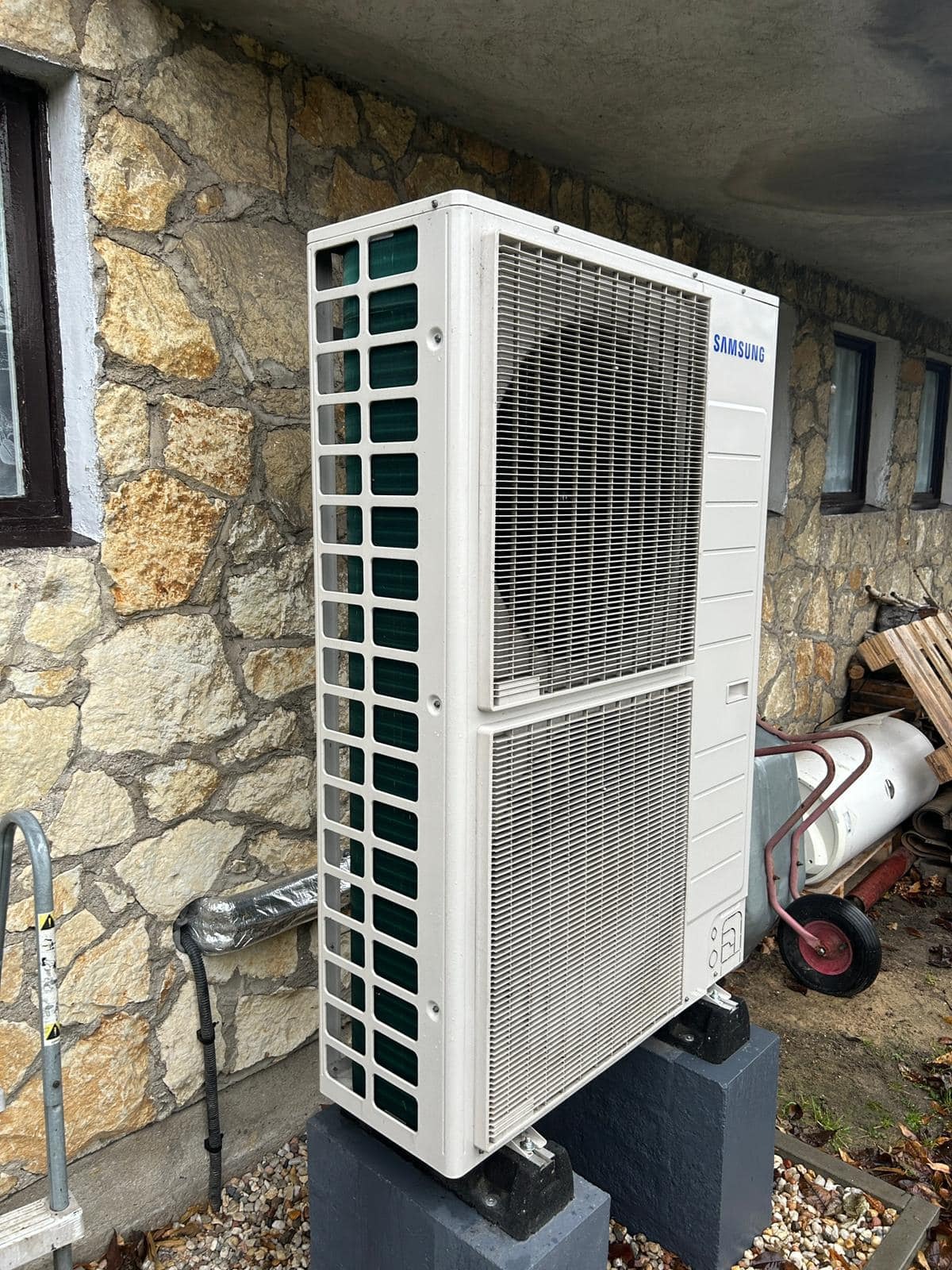 Biała pompa ciepła Samsung na betonowych podstawach, zamontowana przy kamiennej ścianie budynku. Widoczne okna i elementy otoczenia.