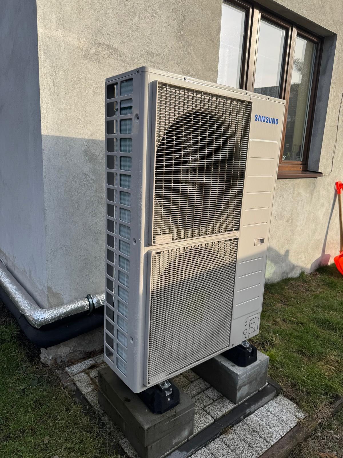 Zewnętrzna jednostka pompy ciepła Samsung, zamontowana na betonowych podstawach przy ścianie budynku z oknem. Widoczne rury w izolacji i trawnik.