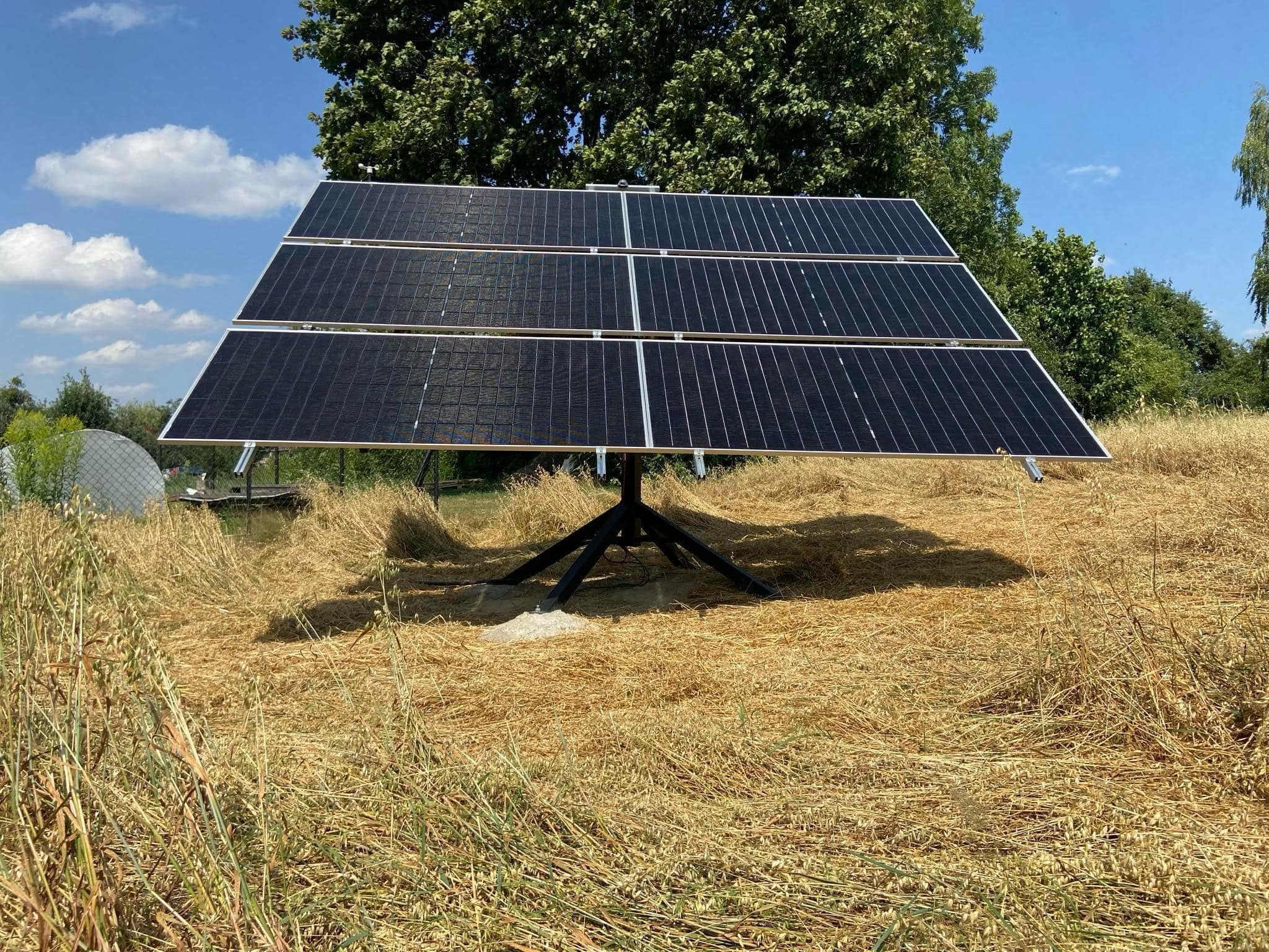 Panel słoneczny na trójnogu, otoczony suchą trawą, z drzewami w tle pod błękitnym niebem. Instalacja fotowoltaiczna w plenerze.