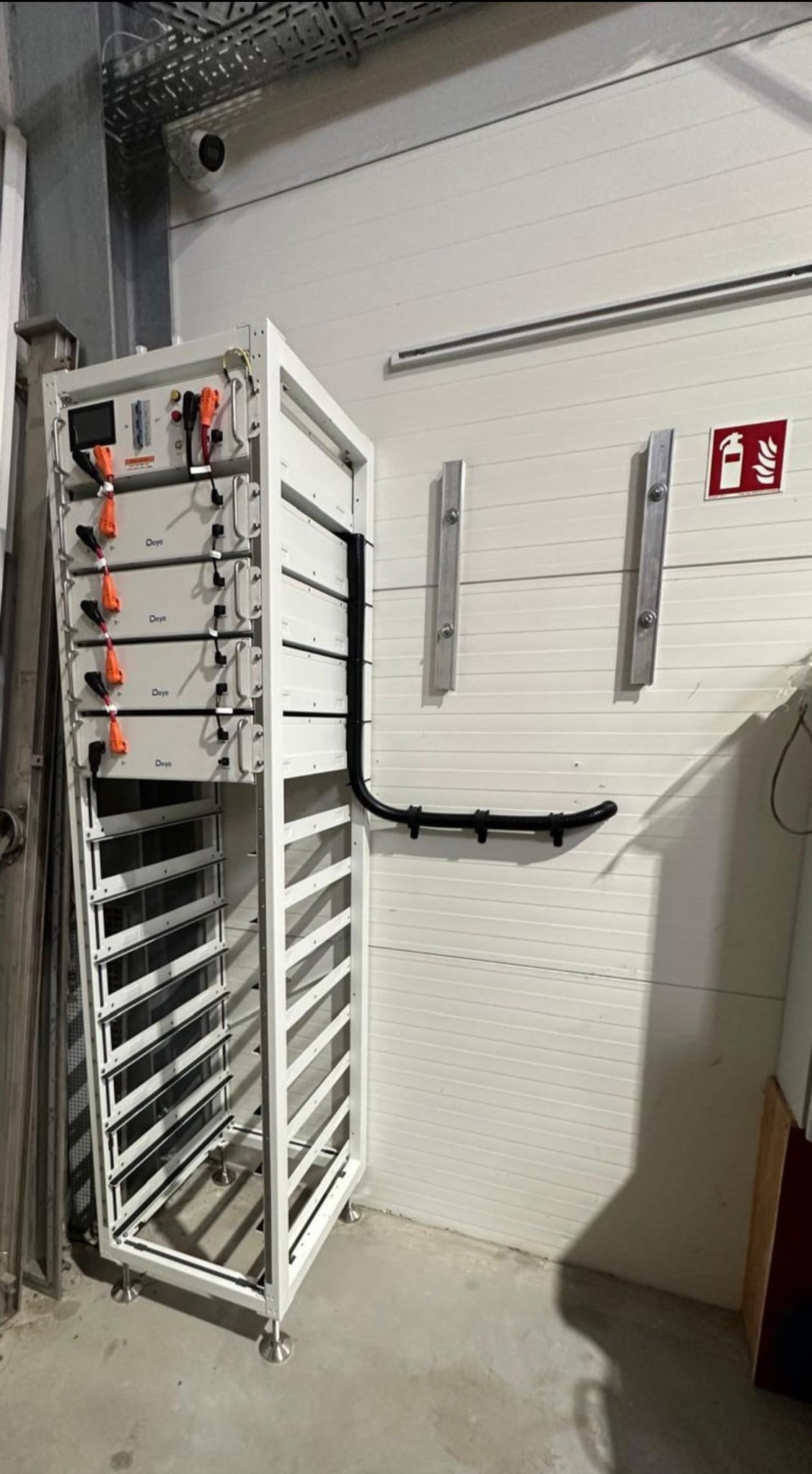 Biała szafa z akumulatorami Deye w hali, widoczne kable i zabezpieczenia. Na ścianie znak gaśnicy i metalowe uchwyty. Instalacja w obiekcie przemysłowym.