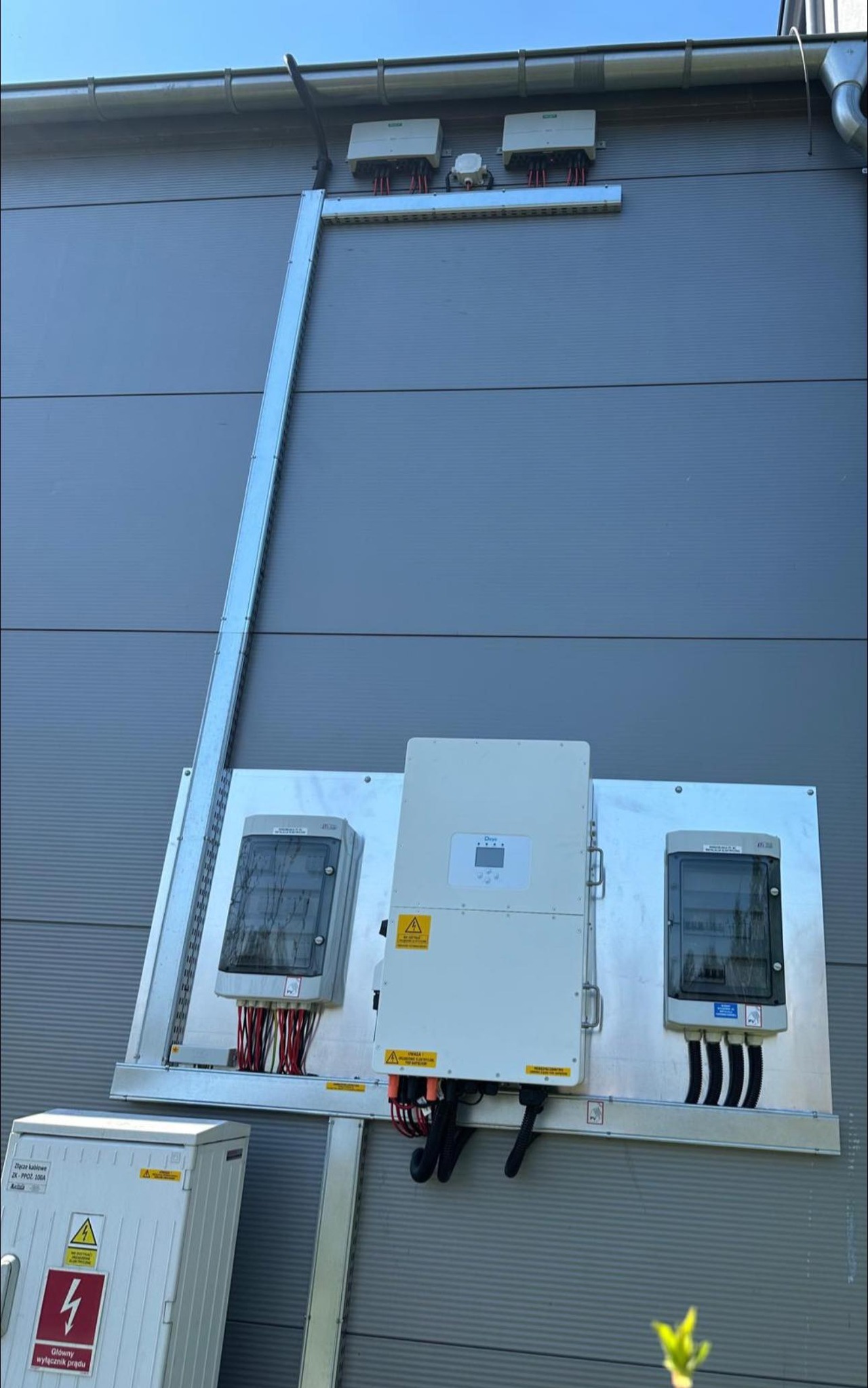 Instalacja paneli fotowoltaicznych na elewacji budynku z widocznymi skrzynkami elektrycznymi i przewodami. Montaż wykonany na metalowej konstrukcji.
