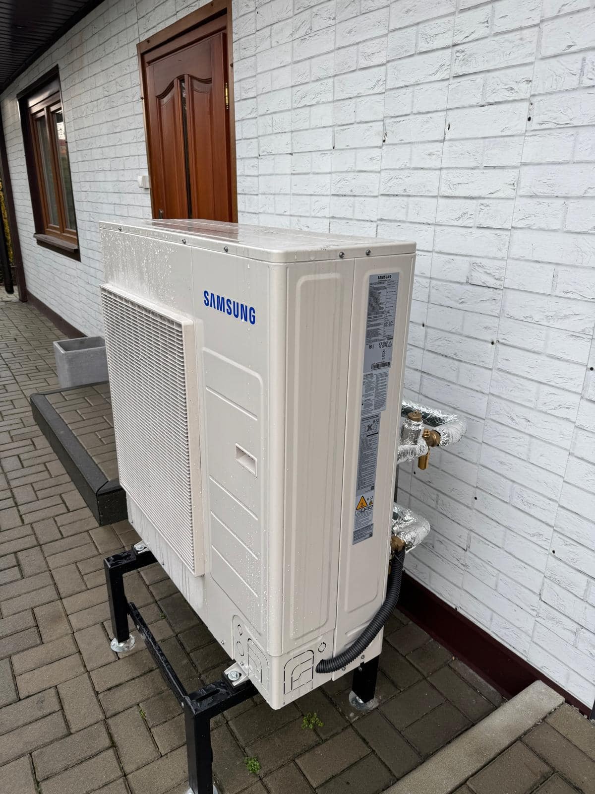 Biała zewnętrzna jednostka pompy ciepła Samsung na czarnym stojaku, przy białej ceglanej ścianie budynku. Widoczne rury w izolacji. Ujęcie z boku, w dzień.