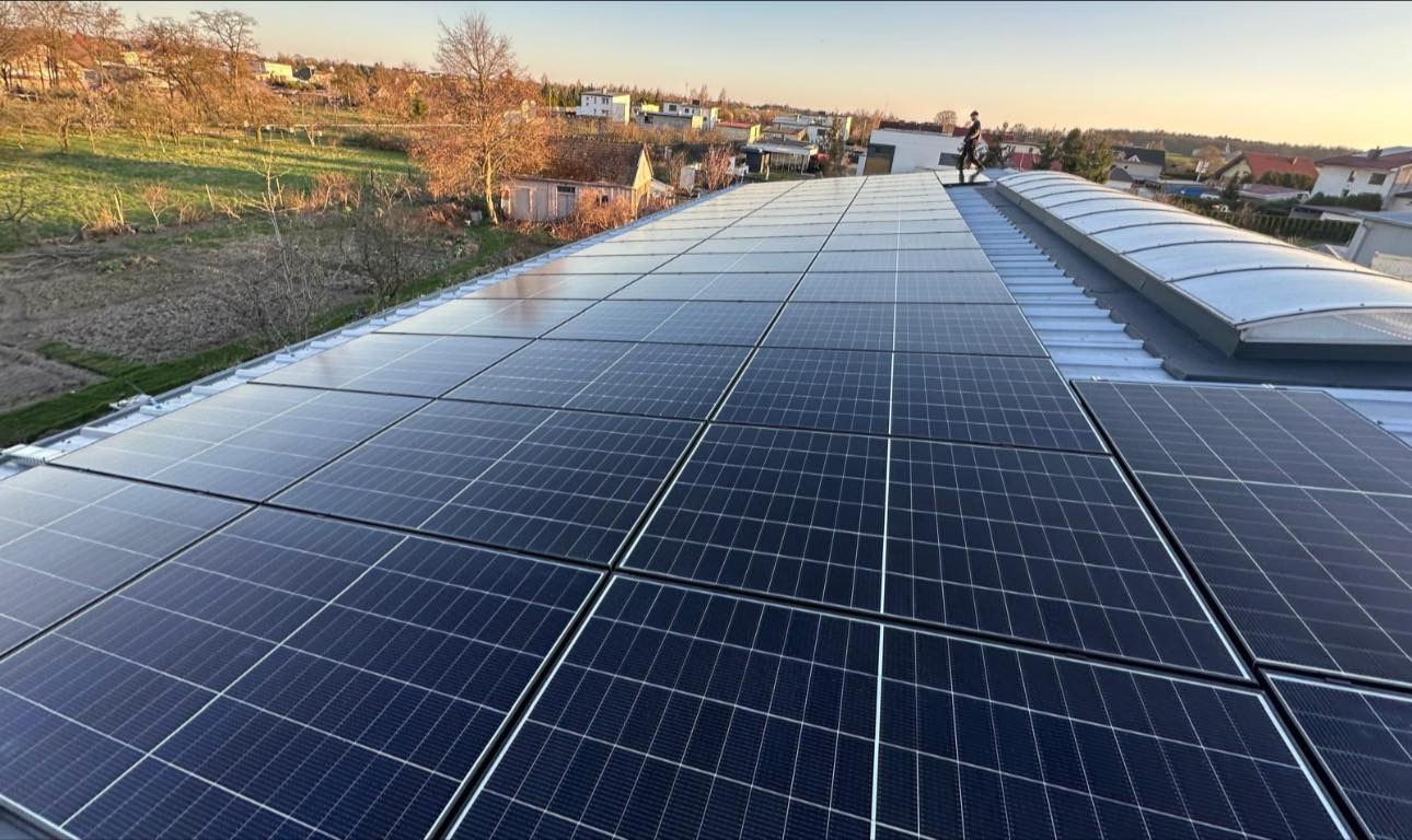 Instalacja paneli słonecznych na dachu budynku mieszkalnego, widok z góry, osoba na dachu, w tle zabudowa jednorodzinna i drzewa, słoneczny dzień.