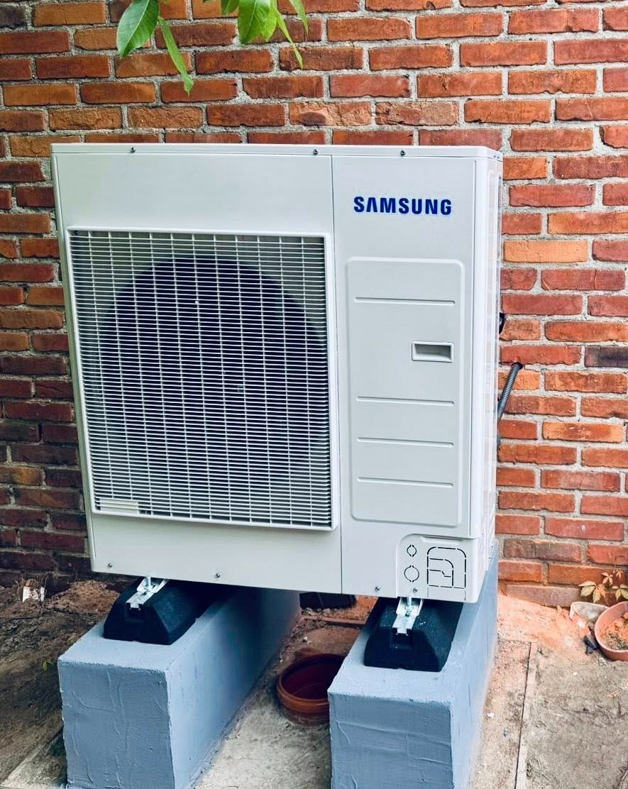 Biała pompa ciepła Samsung na betonowych podstawach, ustawiona przy ceglanej ścianie. Widoczne zielone liście nad urządzeniem i doniczka obok.