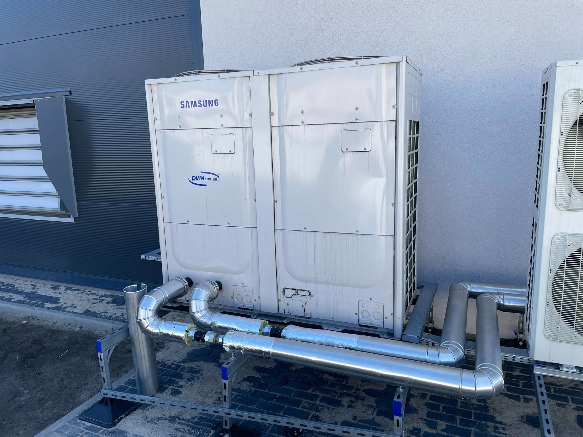 Dwa białe agregaty Samsung DVM Chiller na zewnątrz budynku, z widocznymi srebrnymi rurami instalacji chłodniczej, zamontowane na metalowej konstrukcji wsporczej.
