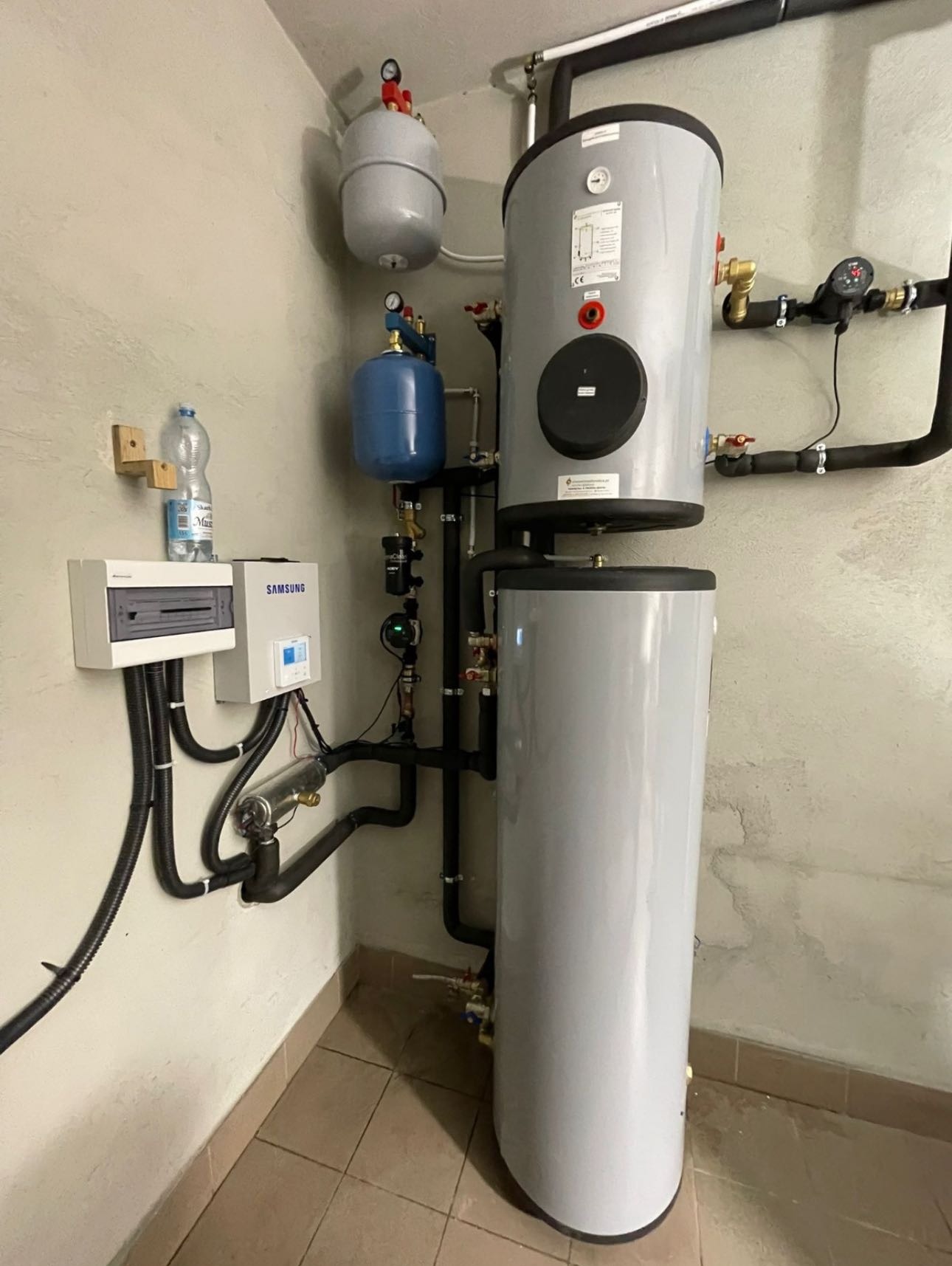 Zestaw instalacji grzewczej z szarym zbiornikiem, rurami i osprzętem na tle beżowej ściany. Widoczna butelka wody na szafce.