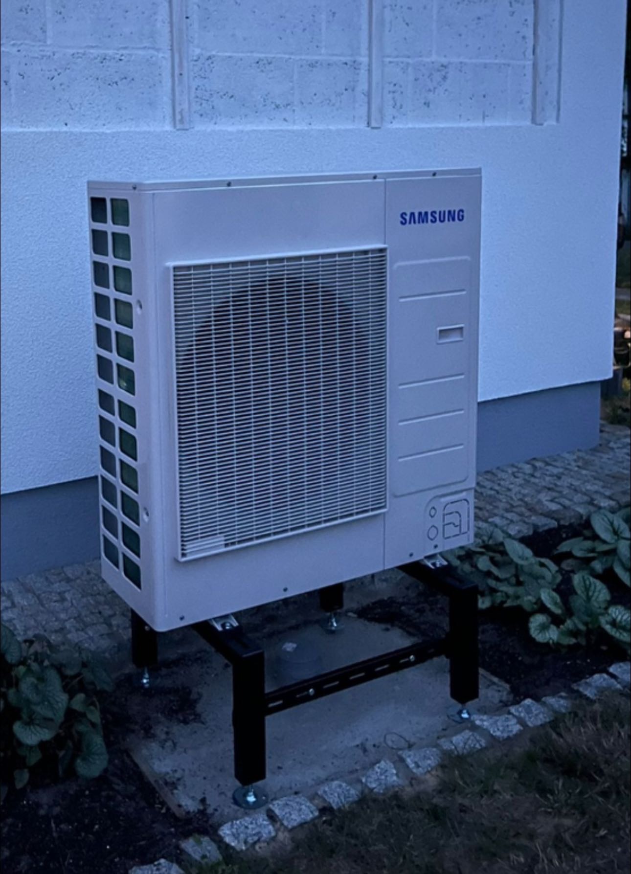 Biała pompa ciepła Samsung na czarnym stelażu, ustawiona przy ścianie budynku z widocznymi elementami instalacji i otoczeniem z roślinnością.