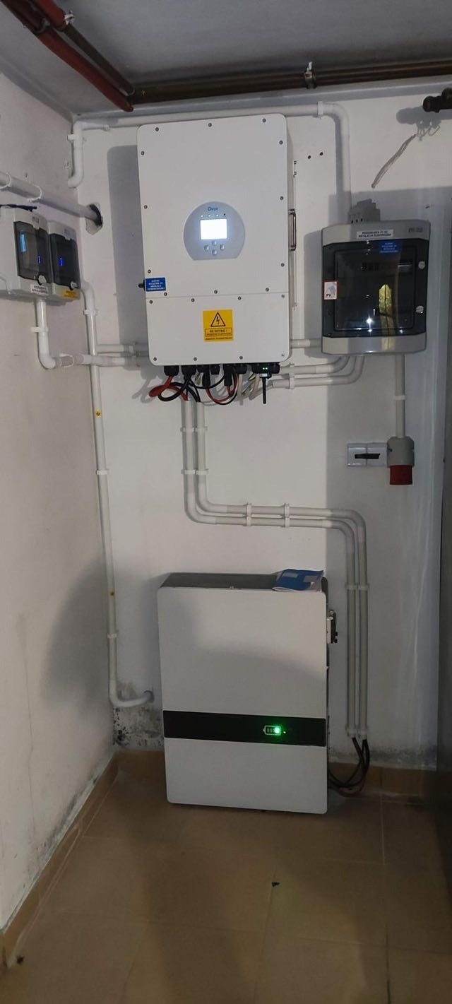 Biała bateria, inwerter i rozdzielnica na ścianie w pomieszczeniu. Widoczne rury instalacyjne i gniazdo elektryczne. Instalacja fotowoltaiczna w piwnicy.