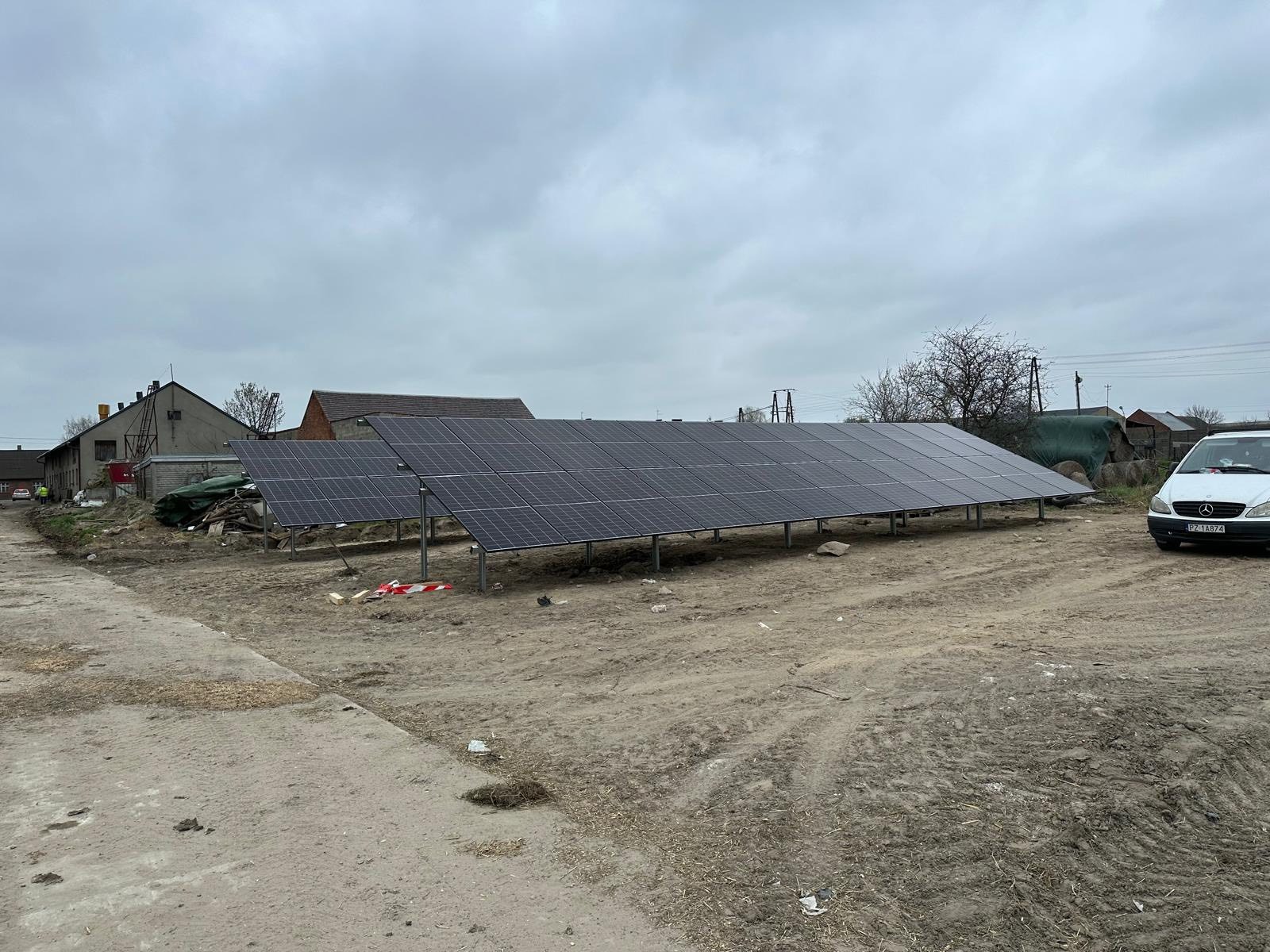 Instalacja paneli fotowoltaicznych na gruncie, widoczne panele w rzędach, teren przygotowany, w tle budynki gospodarcze i zaparkowany biały samochód.