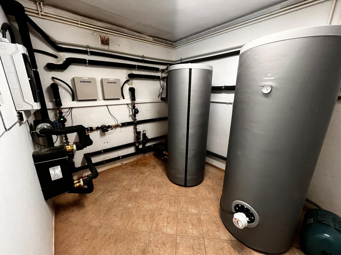 Kompletna instalacja pompy ciepła w kotłowni: dwa zbiorniki Galmet, jednostka Samsung, liczne rury i zawory. Widok na całą instalację w pomieszczeniu.