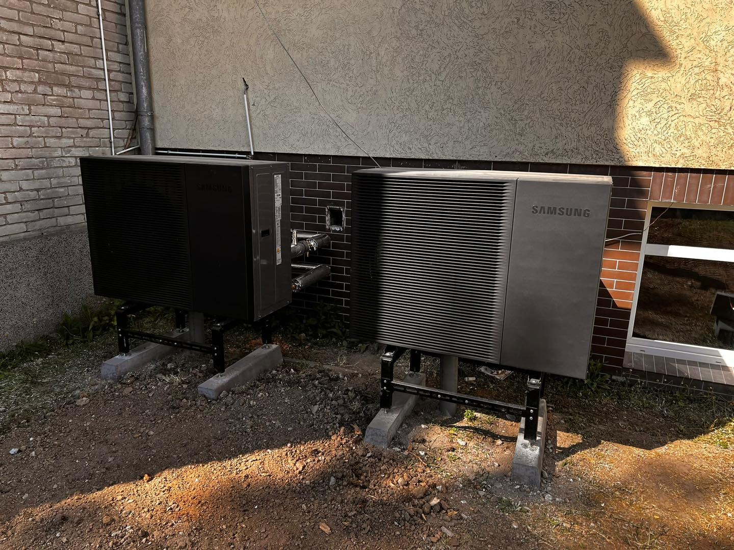 Dwie czarne pompy ciepła Samsung na zewnątrz budynku z ceglaną elewacją i beżowym tynkiem strukturalnym. Widoczne rury i elementy montażowe.