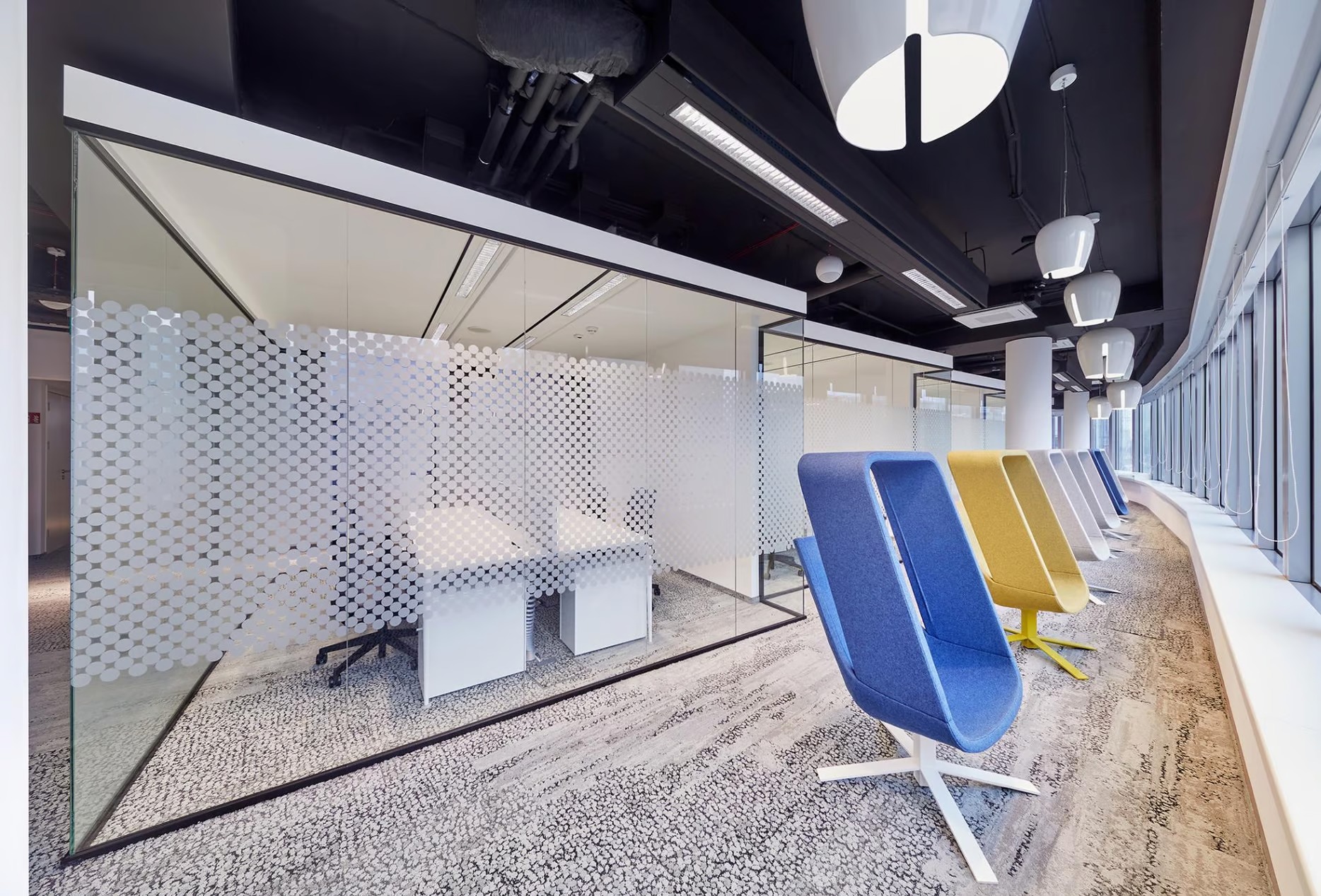 Nowoczesne wnętrze biurowe z przeszklonymi ścianami działowymi z dekoracyjną folią, rzędem kolorowych foteli i designerskimi lampami sufitowymi.