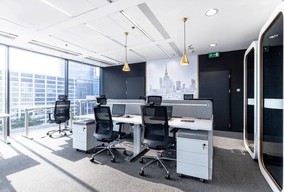 Jasne, nowoczesne biuro z widokiem na miasto, biurka z regulowaną wysokością, ergonomiczne krzesła, kabiny akustyczne i dekoracja ścienna z panoramą Warszawy.