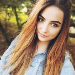 Portret uśmiechniętej kobiety z długimi, rudymi włosami, ubrana w dżinsową kurtkę na tle rozmytej zieleni.