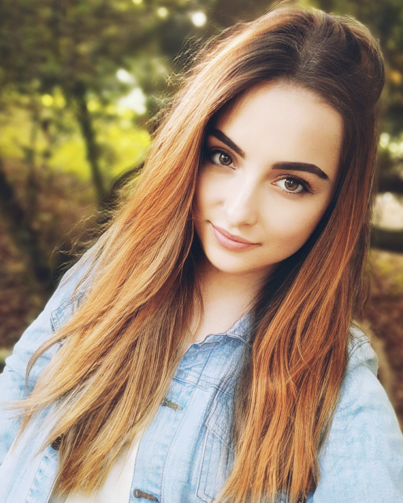 Portret uśmiechniętej kobiety z długimi, rudymi włosami, ubrana w dżinsową kurtkę na tle rozmytej zieleni.