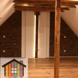 Poddasze po remoncie z drewnianymi belkami stropowymi i kolumnami, ceglana ściana po bokach z białym pasem pośrodku, jasna drewniana podłoga, logo firmy remontowej w lewym dolnym rogu.