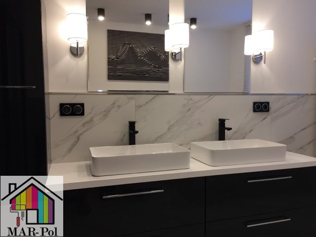 Nowoczesna łazienka z dwoma białymi umywalkami nablatowymi, czarną armaturą, marmurowymi płytkami na ścianie i czarnymi szafkami pod umywalkami, oświetlona kinkietami z białymi abażurami.