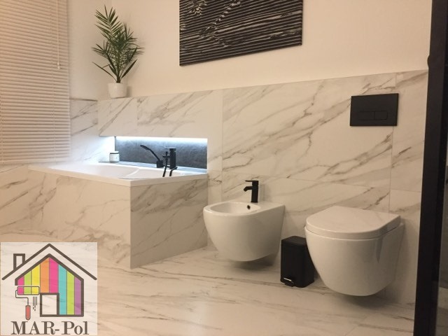 Nowoczesna łazienka z białymi marmurowymi płytkami, podwieszaną toaletą i bidetem, czarną armaturą, oraz rośliną doniczkową nad wanną z podświetleniem LED.