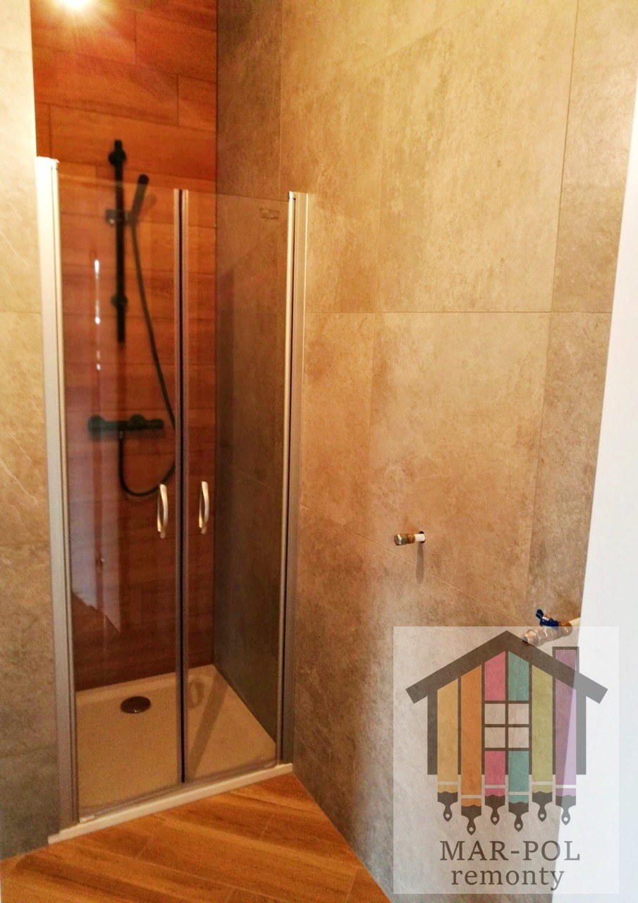 Kabina prysznicowa z przesuwnymi drzwiami w łazience wykończonej płytkami imitującymi drewno, widoczny czarny zestaw natryskowy i logo firmy remontowej w rogu.