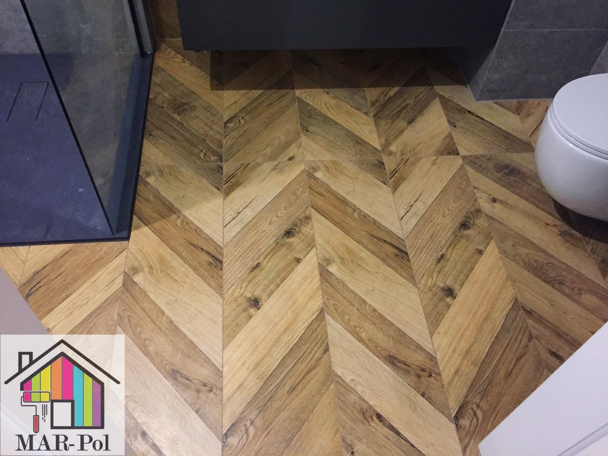 Ułożona pod kątem drewnopodobna podłoga w łazience, widoczny fragment kabiny prysznicowej, toalety i logo firmy w rogu.