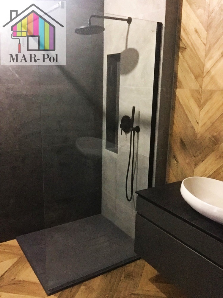 Nowoczesna kabina prysznicowa typu walk-in z czarną armaturą, szarą płytką imitującą kamień i drewnianą podłogą w jodełkę, obok umywalka nablatowa na szafce w tym samym stylu, w lewym górnym rogu...