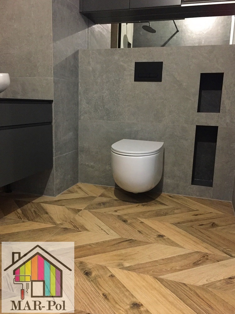 Nowoczesna łazienka z szarą okładziną ścienną, podwieszaną toaletą i drewnopodobną podłogą w jodełkę. Widoczne wnęki w ścianie i logo firmy na podłodze.