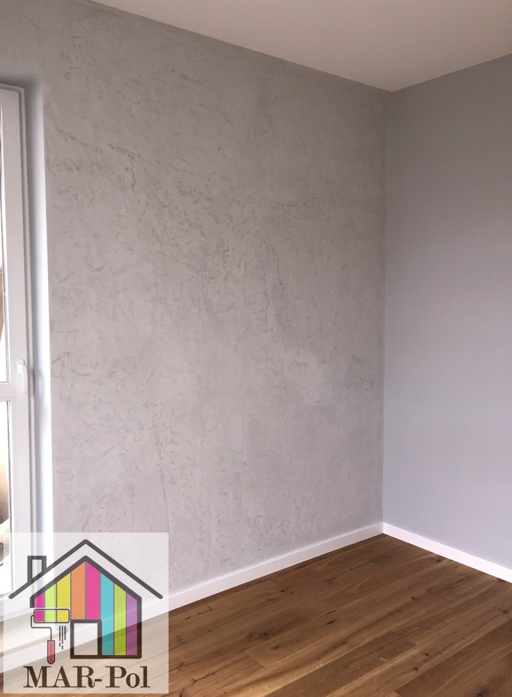 Ściana w pokoju z efektem dekoracyjnym w odcieniach szarości, widoczna faktura na ścianie, drewniana podłoga, biała listwa przypodłogowa, okno po lewej stronie, w rogu logo firmy MAR-Pol.