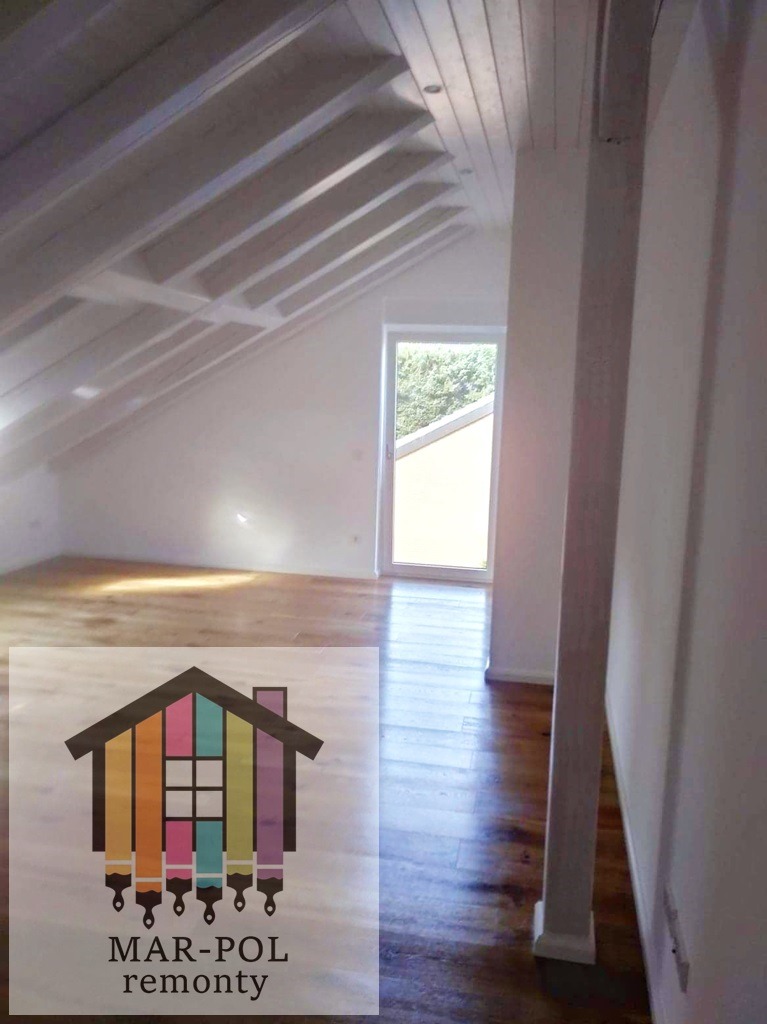 Poddasze po remoncie z drewnianą podłogą, białe ściany i sufit z widocznymi belkami, okno w szczycie, w rogu widoczny słup nośny, na pierwszym planie logo firmy remontowej.
