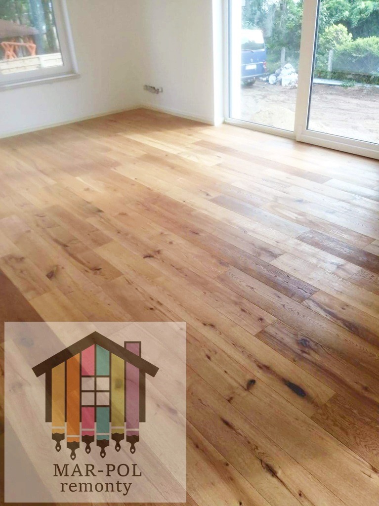 Ułożona pod kątem jasna drewniana podłoga w pustym pokoju z dużym oknem balkonowym, widoczne logo firmy MAR-POL remonty.