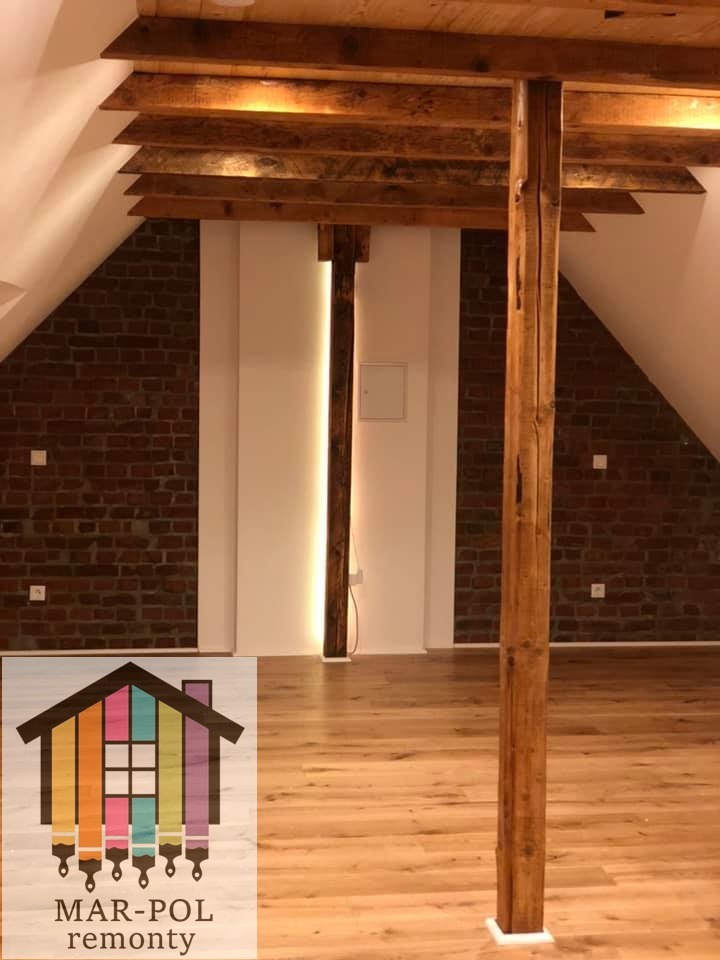 Poddasze po remoncie z drewnianymi belkami stropowymi i kolumnami, ceglana ściana po bokach z białym pasem pośrodku, jasna drewniana podłoga, logo firmy remontowej w lewym dolnym rogu.