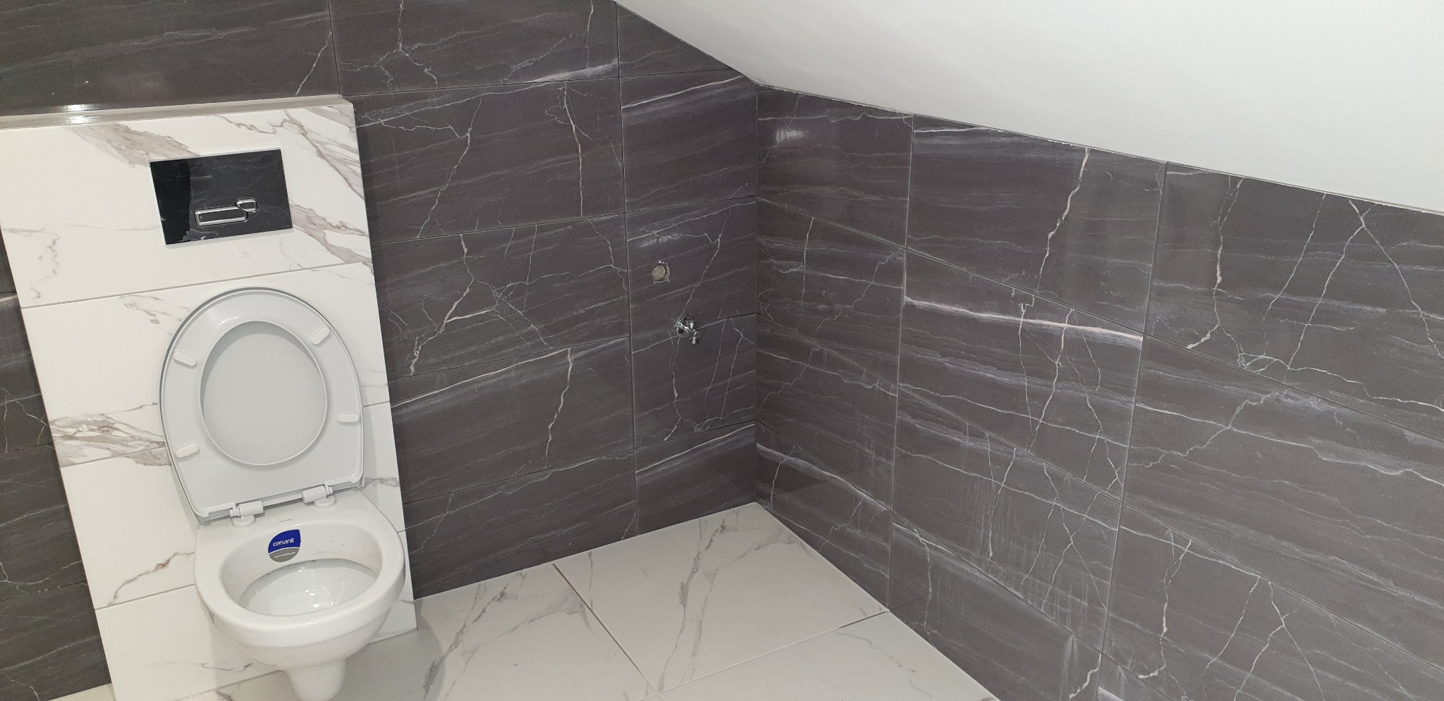 Wykończona łazienka na poddaszu z toaletą podwieszaną i szarymi płytkami imitującymi marmur na ścianach, oraz białymi płytkami na podłodze.