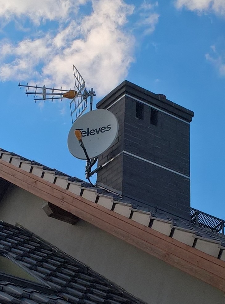 Antena satelitarna marki Televes i antena kierunkowa UHF zamontowane na kominie domu z ciemnej dachówki, błękitne niebo z chmurami w tle.
