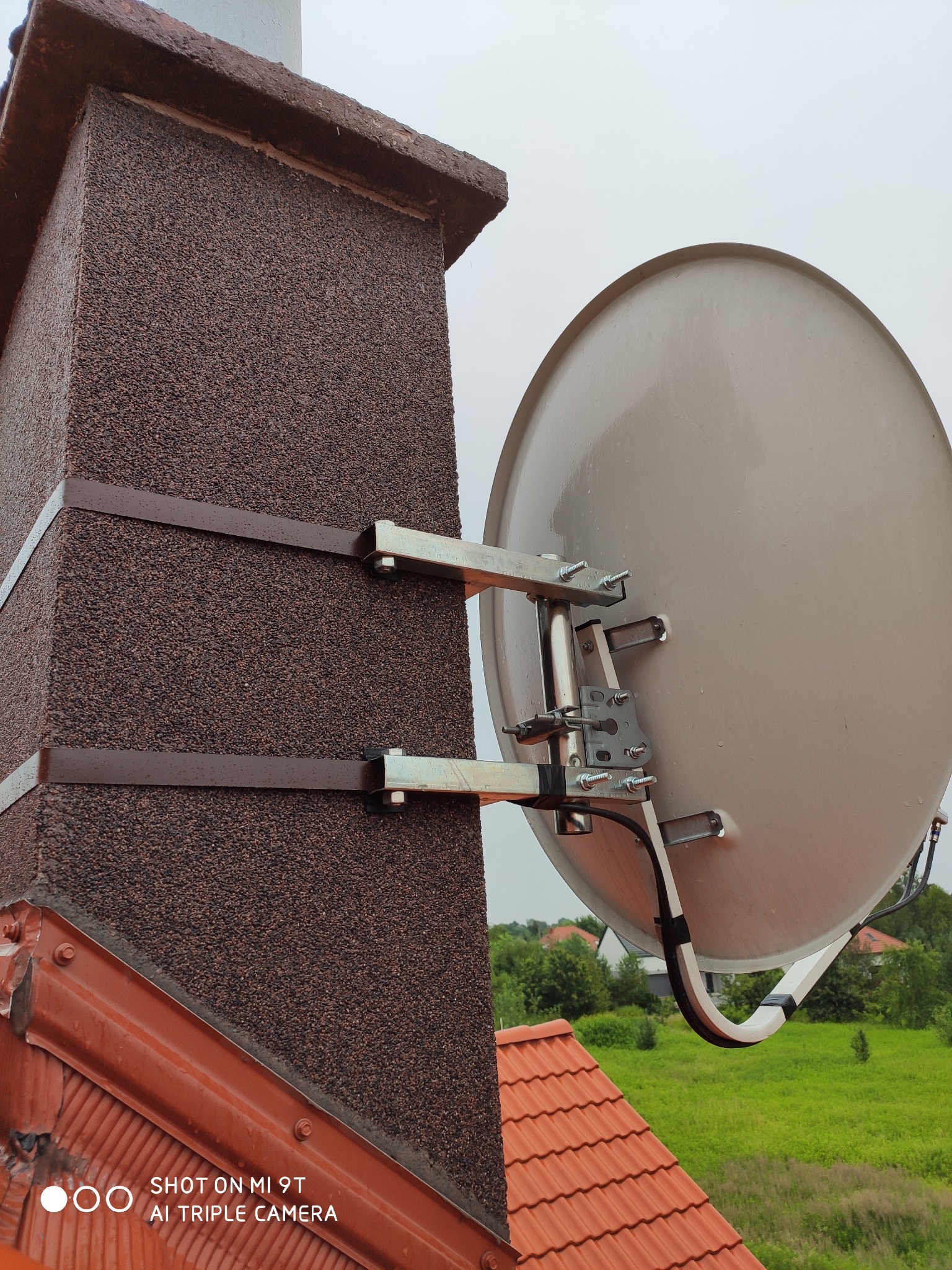 Antena satelitarna zamontowana na kominie z brązowym tynkiem strukturalnym, przymocowana za pomocą metalowych obejm i wsporników, widok z dachu pokrytego czerwoną dachówką, w tle zieleń drzew...