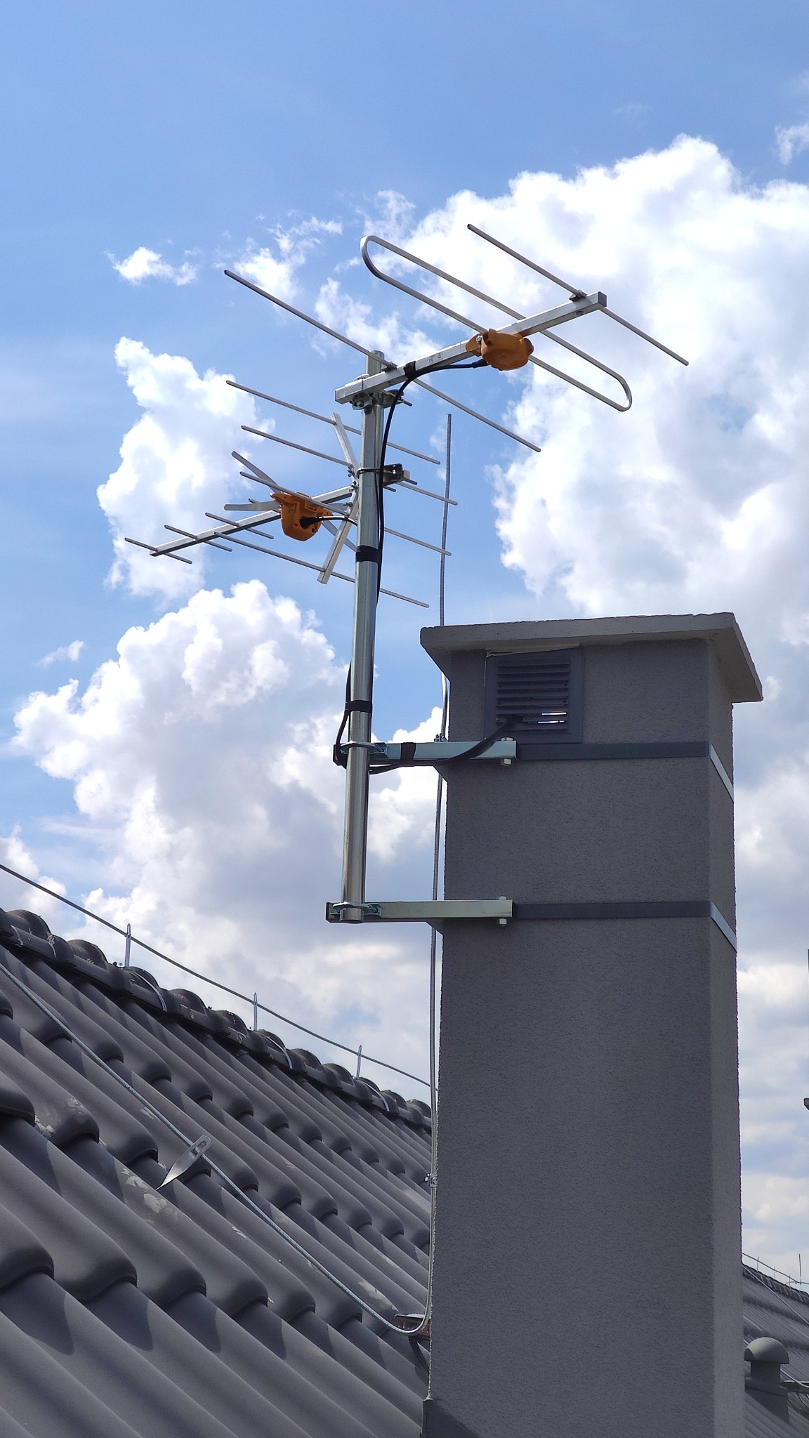 Antena telewizyjna zamontowana na kominie, widok z dołu na tle błękitnego nieba z chmurami, widoczne elementy mocujące do komina i dachu.