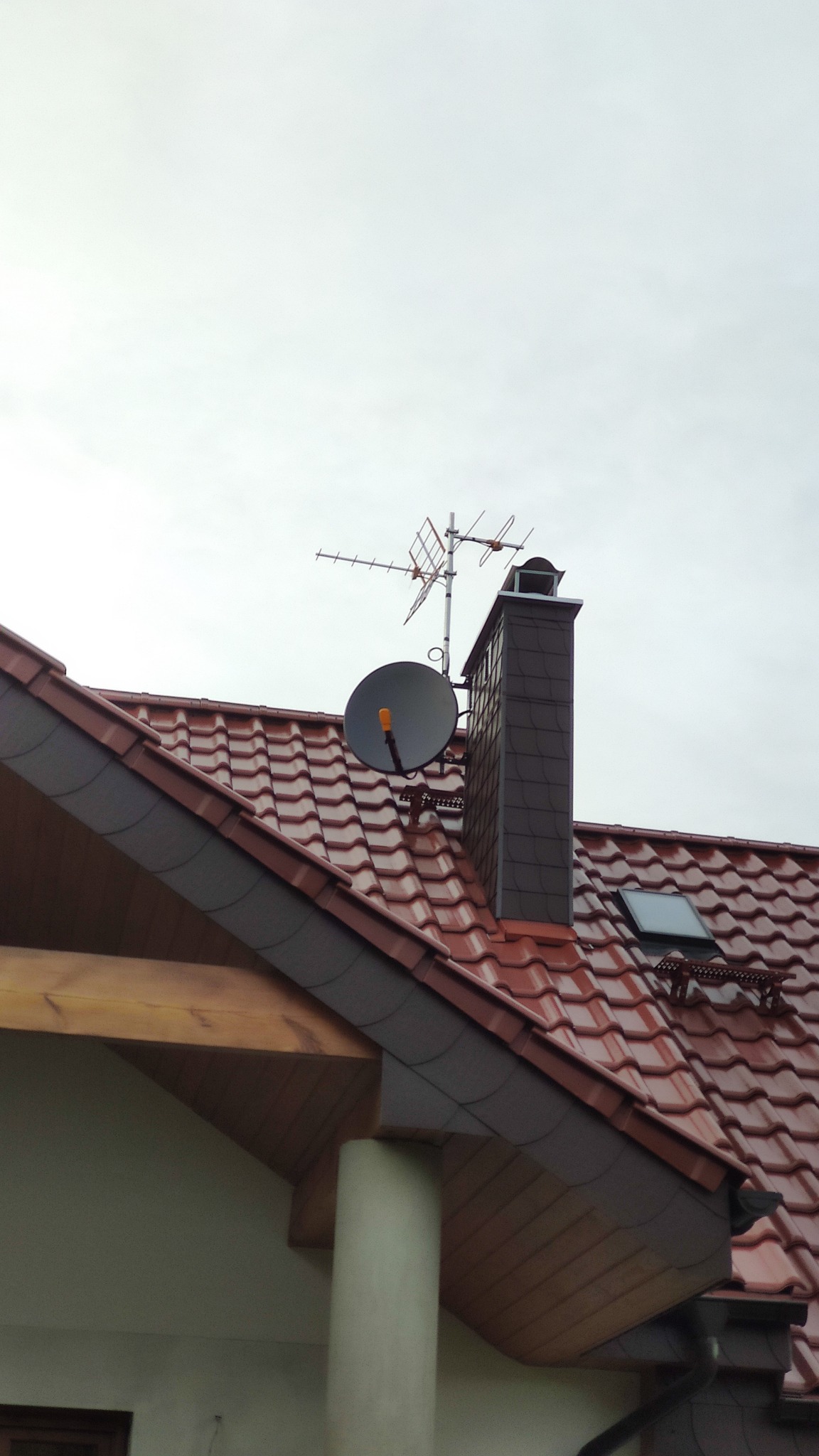 Antena satelitarna i antena naziemna zamontowane na brązowym dachu z dachówką ceramiczną, obok komina z szarej cegły.