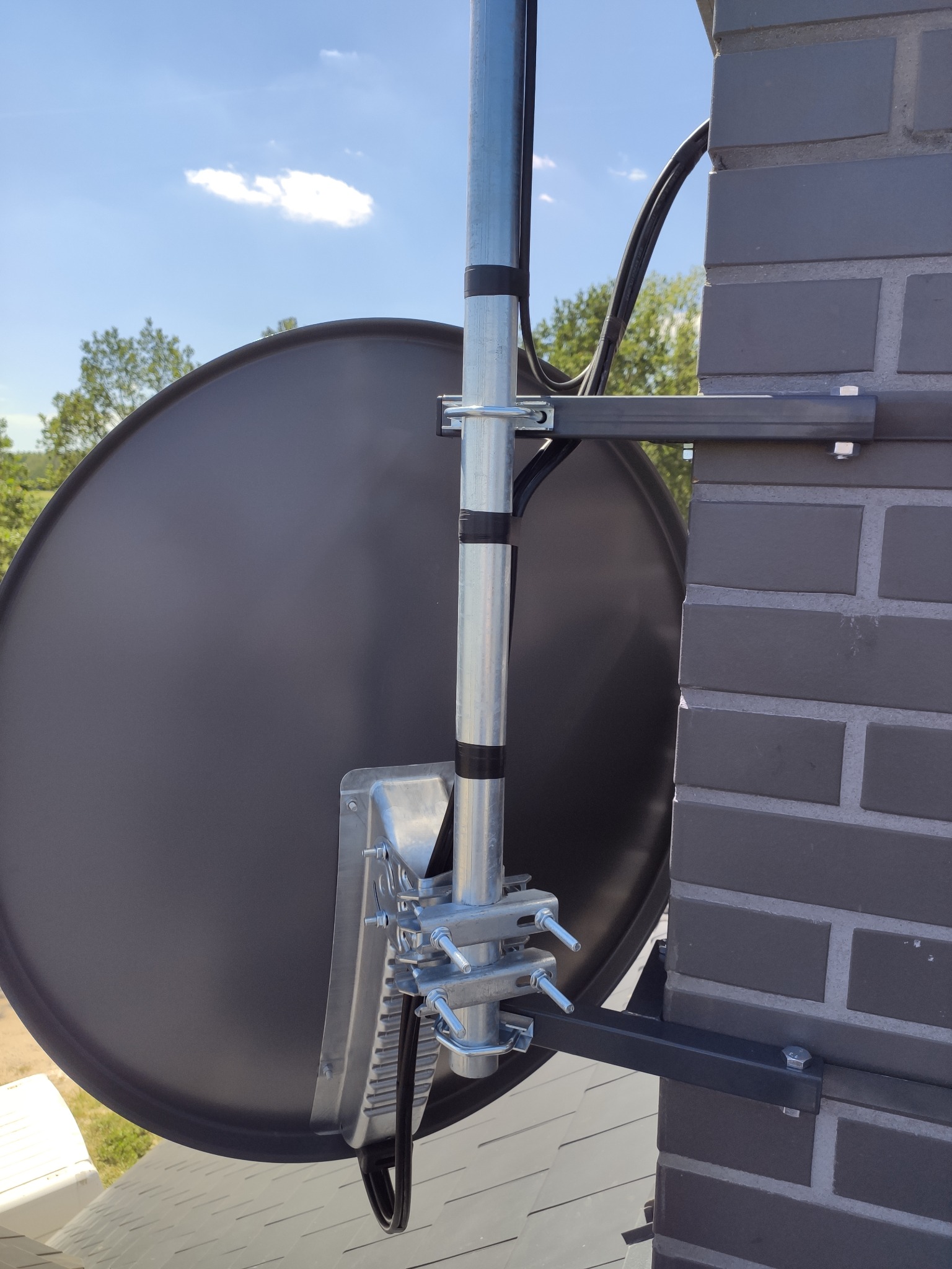 Czarna antena satelitarna zamocowana do komina z szarej cegły, widok z dołu na tle błękitnego nieba z chmurami.