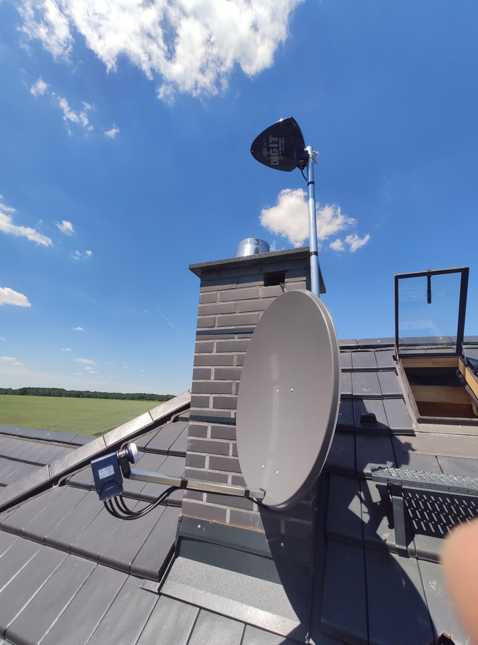 Antena satelitarna i antena DVB-T zamontowane na dachu z szarych dachówek, widok na komin z cegły klinkierowej i otwarte okno dachowe, w tle zielone pole i błękitne niebo z chmurami.